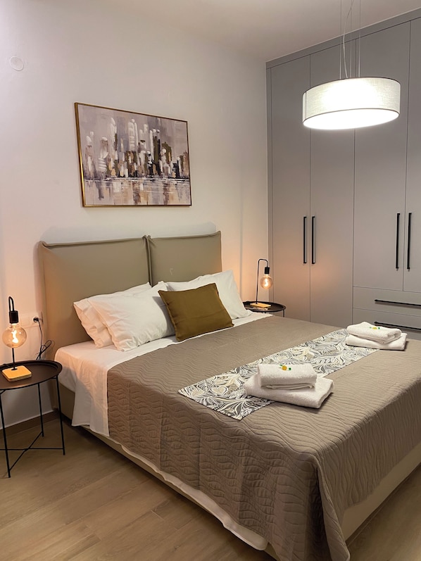 A neatly made bed with a quilt, pillows, and folded towels. There is a bedside table with a lamp, a wall-mounted painting, and a wardrobe.