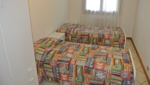 A small room with two beds, each with a colorful quilt. There is a window with sheer curtains and a white radiator on the wall.