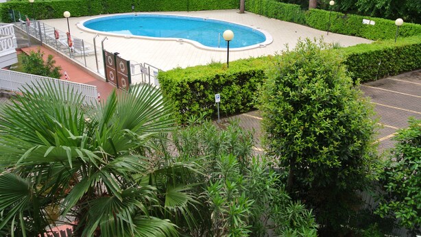 A swimming pool surrounded by well-maintained hedges and palm trees, with a parking area in the foreground.