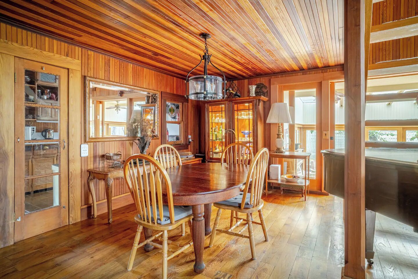 Uma sala de jantar com painéis de madeira, uma grande mesa redonda e cadeiras, um candelabro e vista para o exterior.