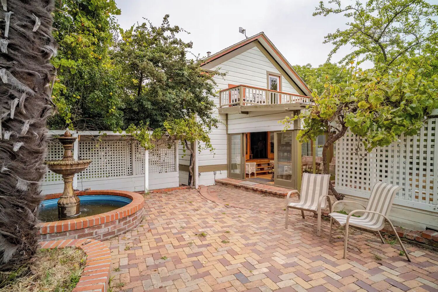 Un patio trasero con un adoquín, una fuente, una cerca blanca y una casa de dos pisos con un balcón.