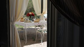 Un área de comedor al aire libre con una mesa preparada para dos personas, rodeada de sillas blancas, bajo una estructura cubierta con cortinas blancas.