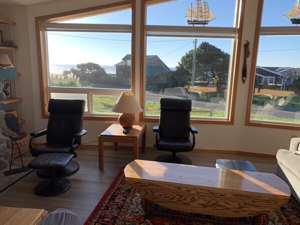 A living room with two black chairs, a wooden coffee table, and a lamp. Large windows offer a view of the sea and houses.