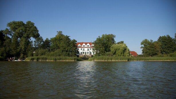 Una casa junto a un lago tranquilo con techo rojo, rodeada de árboles.