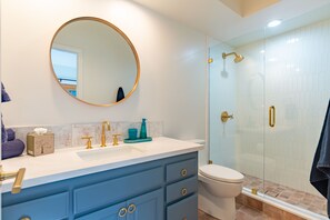 A bathroom with a round mirror, a white sink, a blue vanity, a toilet, and a glass-enclosed shower area. - Serene Garden Retreat in a Charming Rancho Santa Fe Guest House (Rancho Santa Fe)