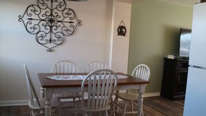 A dining area with a wooden table and white chairs, a decorative wall piece, a small hanging ornament, and a refrigerator in the background. - Cozy Renovated Condo (Miles City)