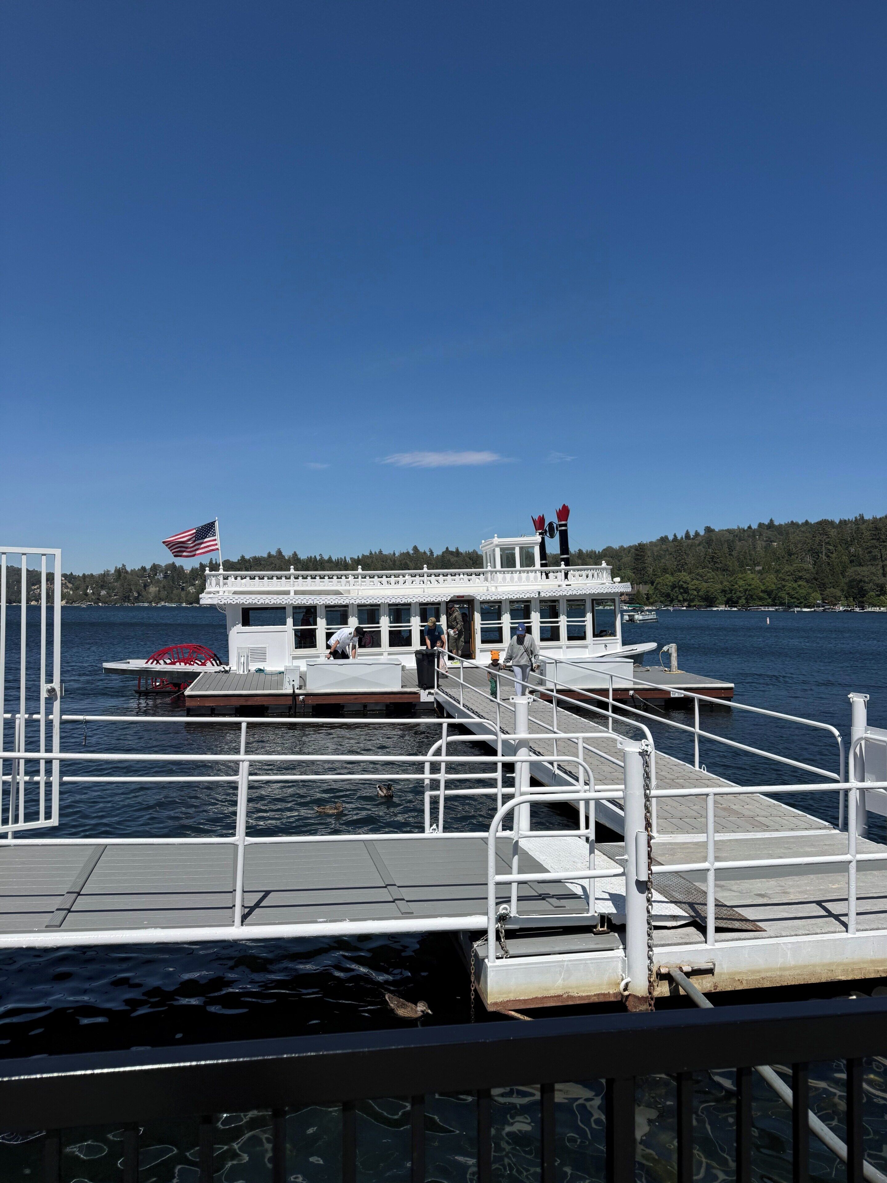 A boat docked at a pier with a flag, a small building, and a forested hill in the background.