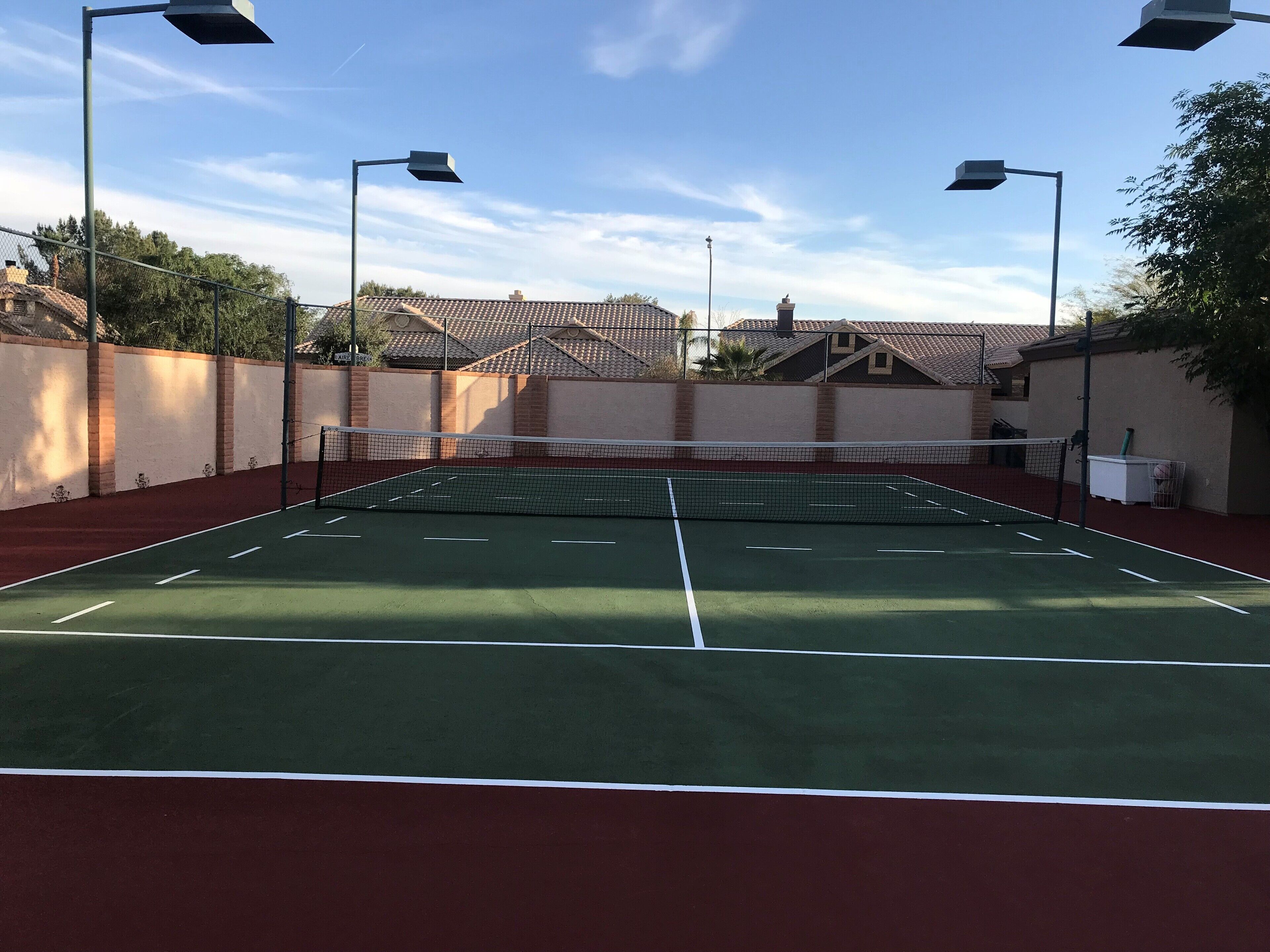 An outdoor tennis court with a net, surrounded by walls and street lamps.