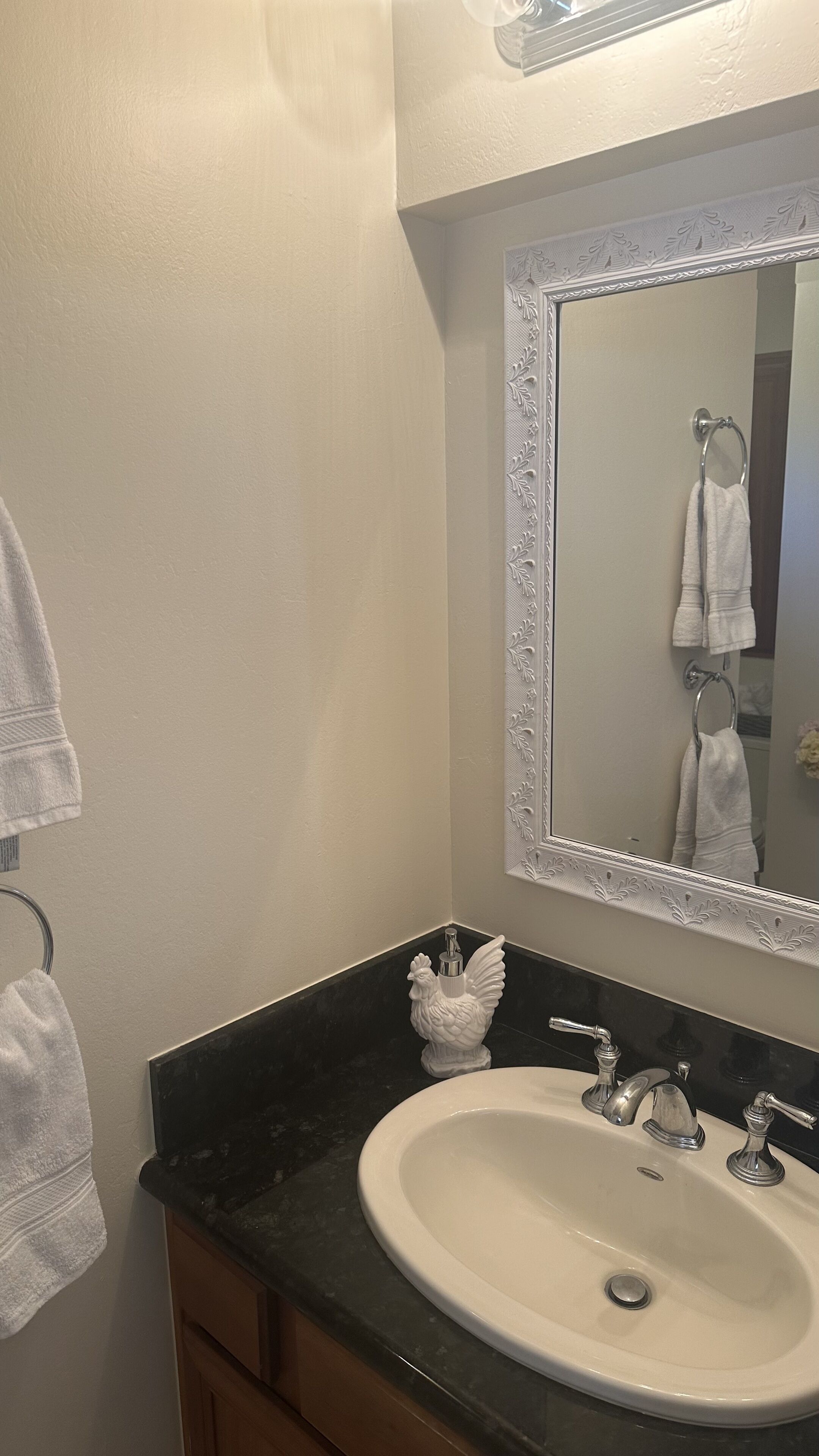 Une salle de bain avec un lavabo blanc, un plan de toilette noir et un miroir doté d’un cadre décoratif. Deux serviettes sont accrochées à des crochets.