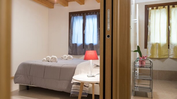 A bedroom with a bed, a bedside table with a lamp, a small chair, a metal shelf with items, and a window with curtains. - Accogliente alloggio vicino al centro (Verona)