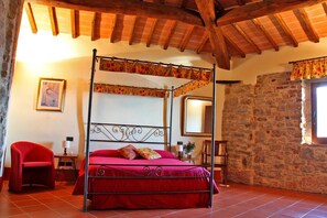 Un lit à baldaquin orné d’un motif floral, un mur en pierre et un fauteuil rouge dans une pièce rustique aux poutres apparentes.