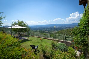 Un jardín con un pabellón, una mesa y sillas, rodeado de exuberante vegetación y vistas a colinas distantes.
