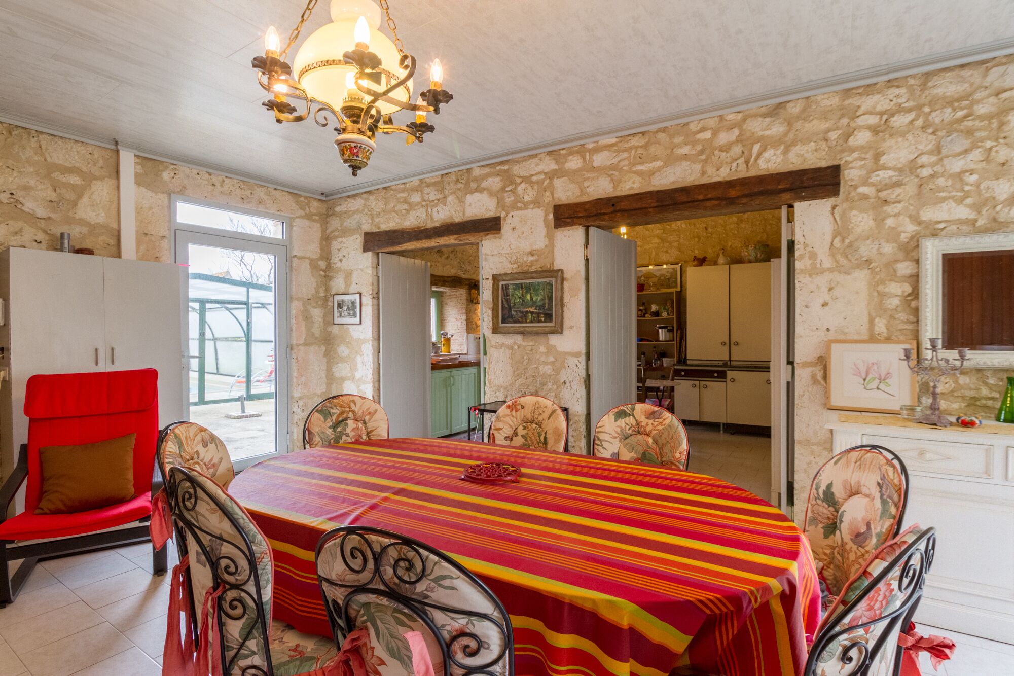 Un coin repas avec une table ronde recouverte d’une nappe à rayures, entourée de chaises à motifs floraux. Un lustre est suspendu au plafond, et la pièce est dotée de murs en pierre et de poutres en bois.