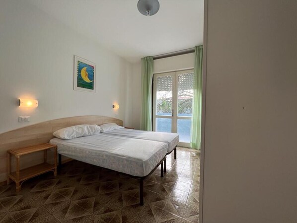 Une chambre à coucher avec un lit, une table de chevet, une lampe fixée au mur, un tableau et une fenêtre avec des rideaux.