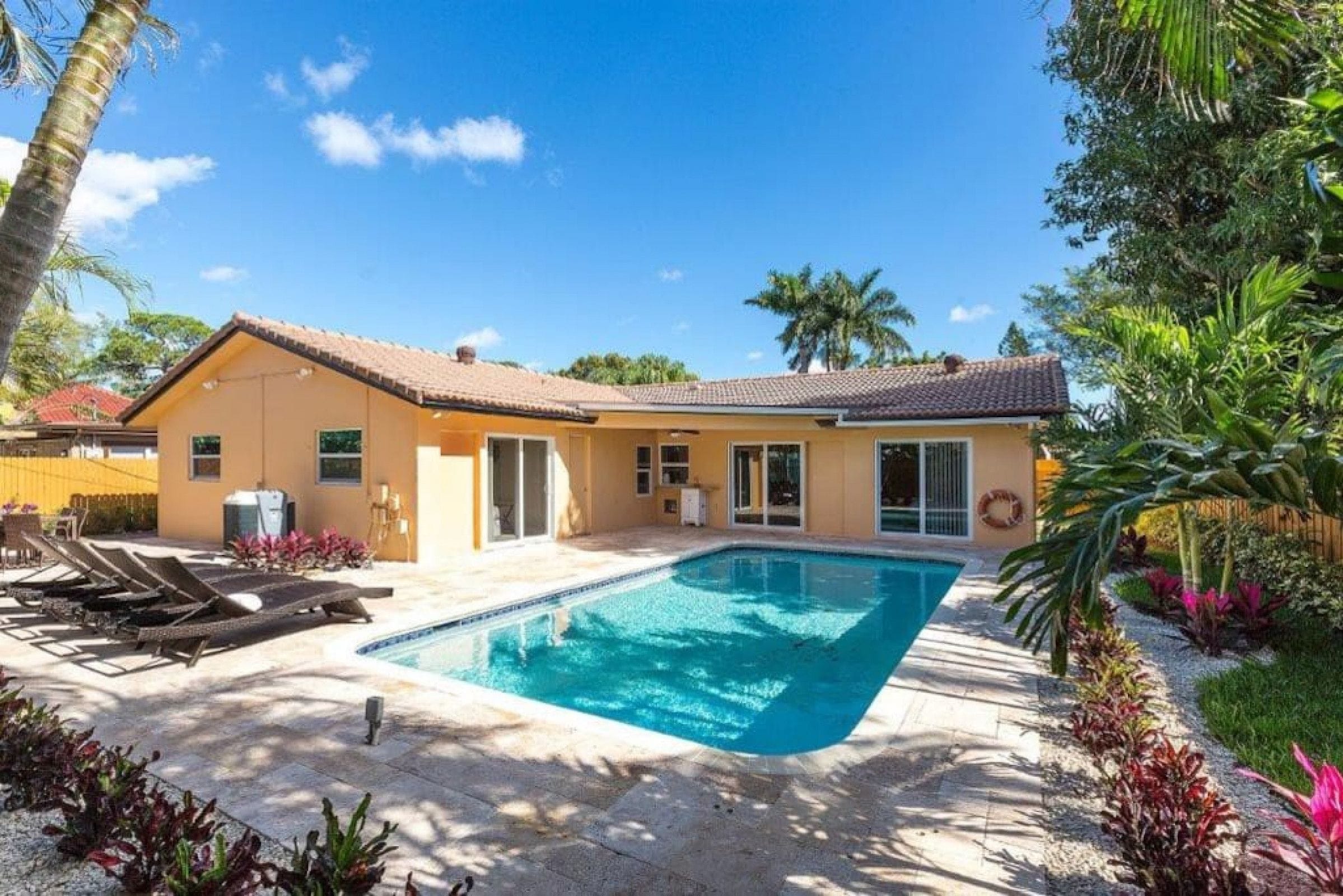 A house with a swimming pool, a patio with lounge chairs, and a garden with tropical plants.