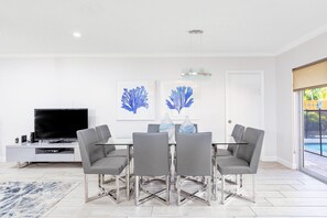 Un espace repas moderne avec une table en verre, des chaises grises et un téléviseur à écran plat posé sur une console blanche.
