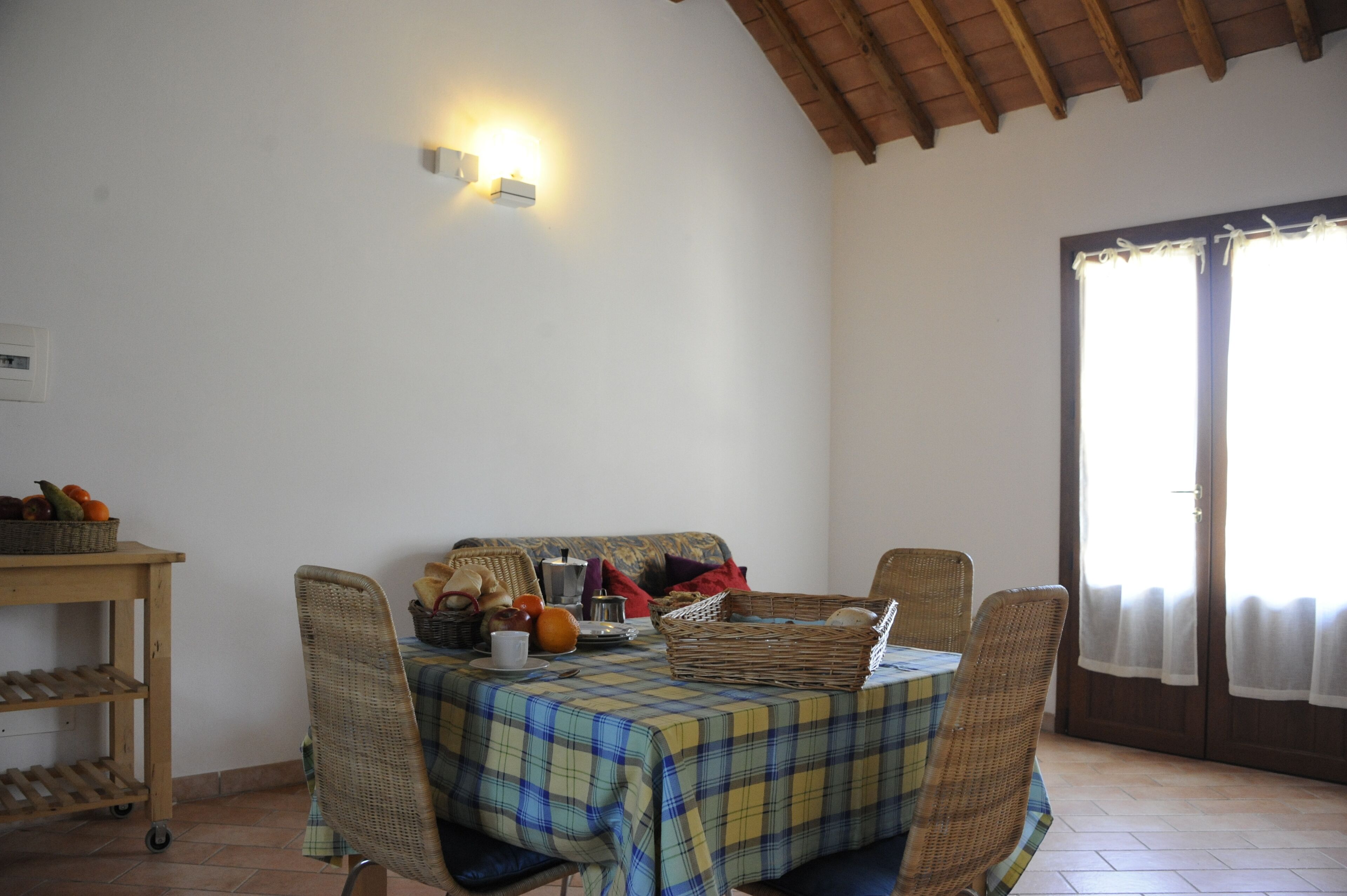 Un comedor con una mesa lista para cuatro personas, rodeada de sillas de mimbre, una cesta de mimbre y una silla de mimbre con cojín.