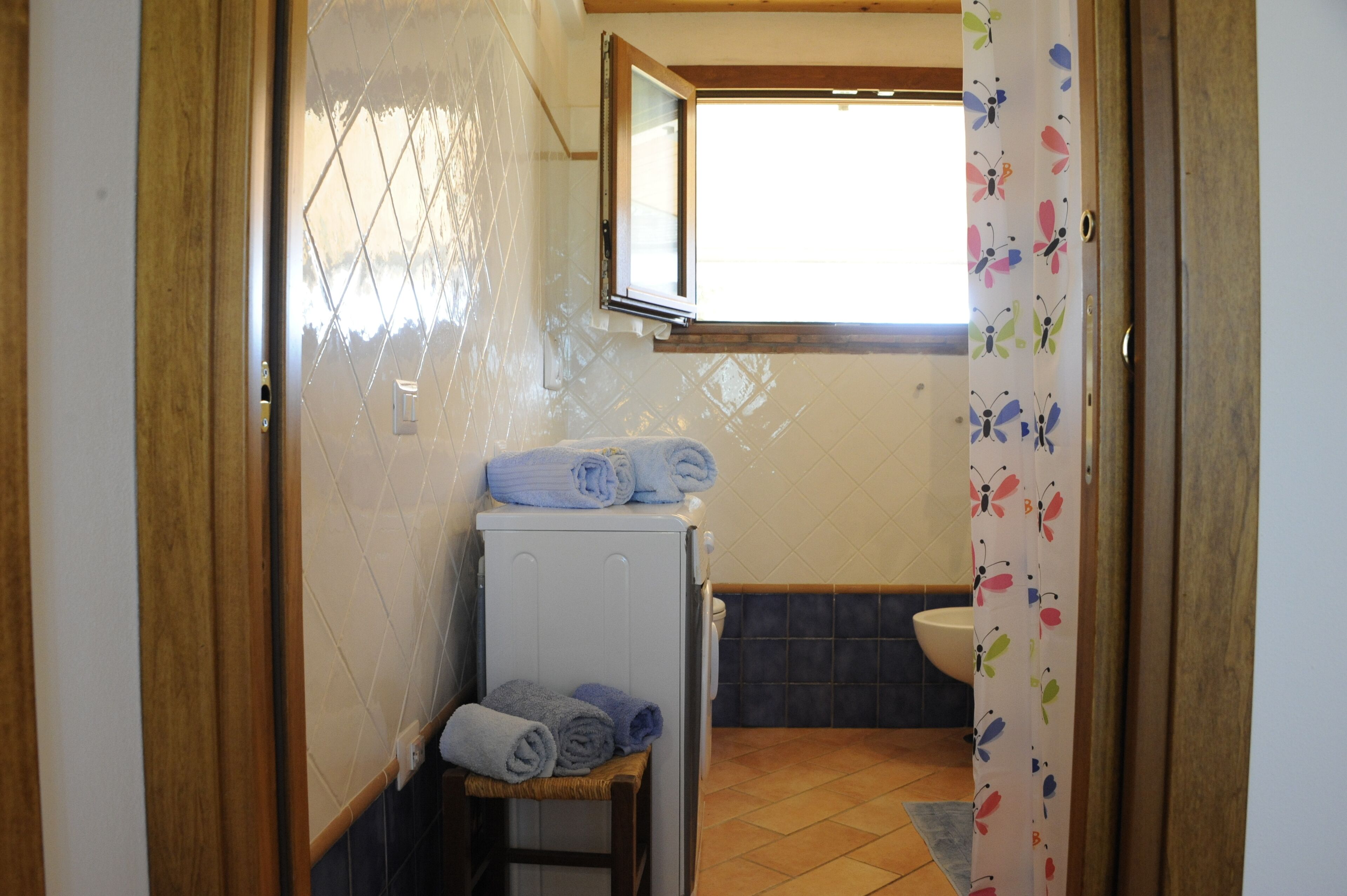 Een kleine badkamer met een witte wasmachine, een houten kruk met handdoeken, een raam en een toilet.
