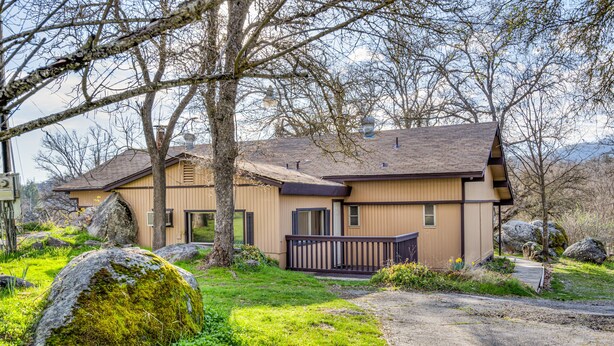 Une maison à deux étages avec une terrasse en bois, de gros rochers et une cour gazonnée.