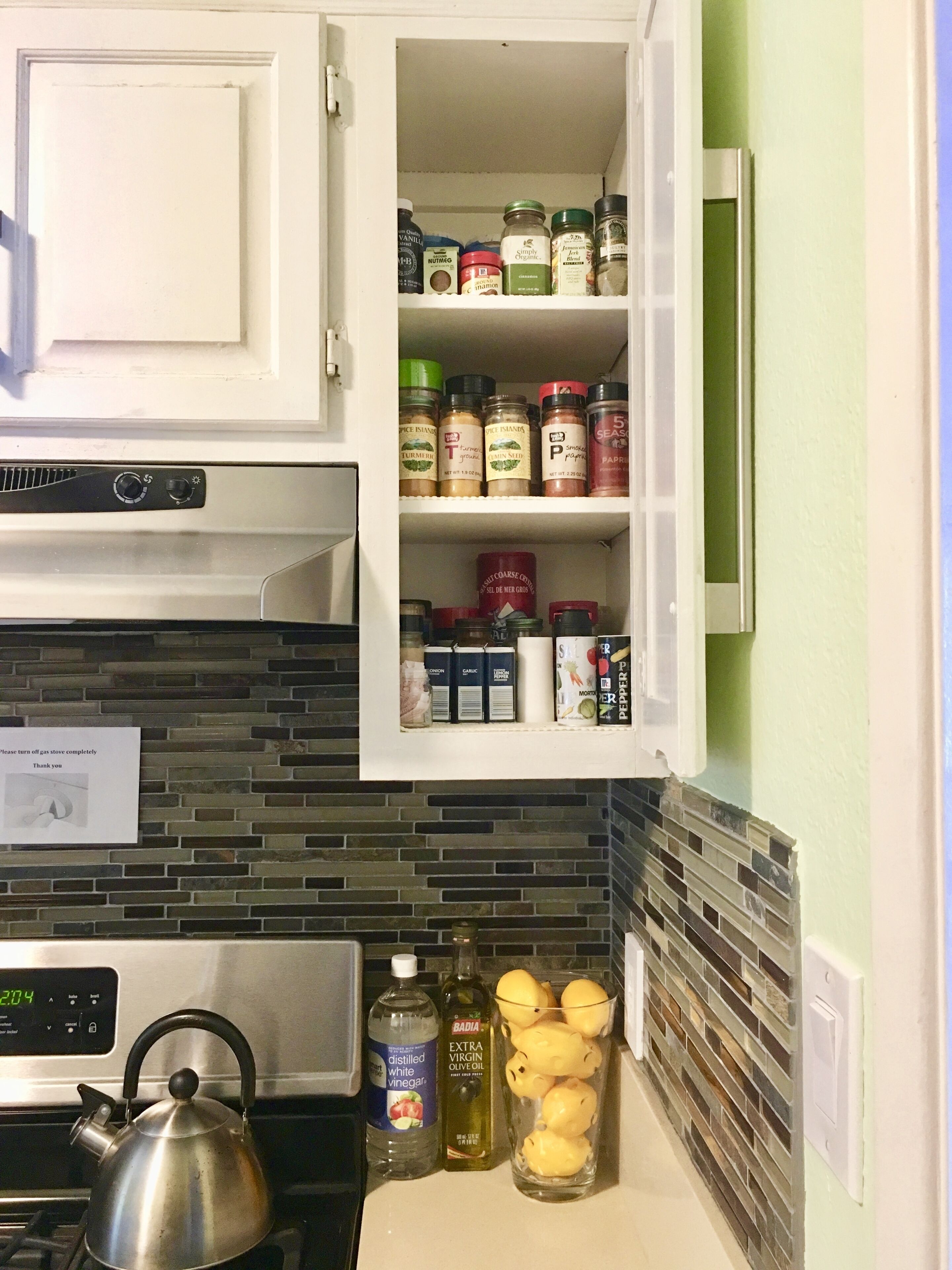 A kitchen with a stove, kettle, and various spices and condiments on the shelves.