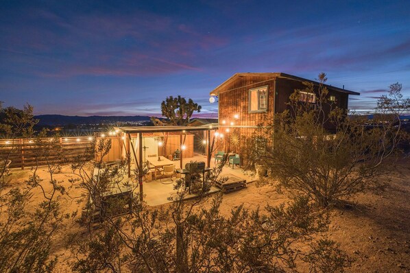 A modern wooden house with a covered patio area, surrounded by desert vegetation.