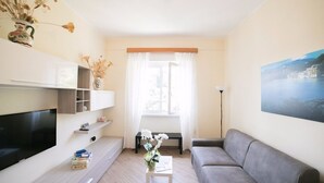 En stue med et fladskærms-tv, en grå sofa, et lille sofabord med en vase og et billede på væggen.