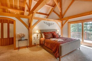 A bedroom with a large bed, a nightstand with a lamp, a wooden ceiling, and a view of the outdoors. - Snowgrass Lodge - A River & Mountain Retreat (Leavenworth)