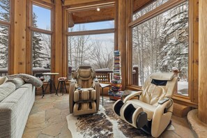 Une pièce chaleureuse avec un fauteuil de massage, une pile de livres et une vue sur une forêt enneigée, visible à travers de grandes fenêtres.