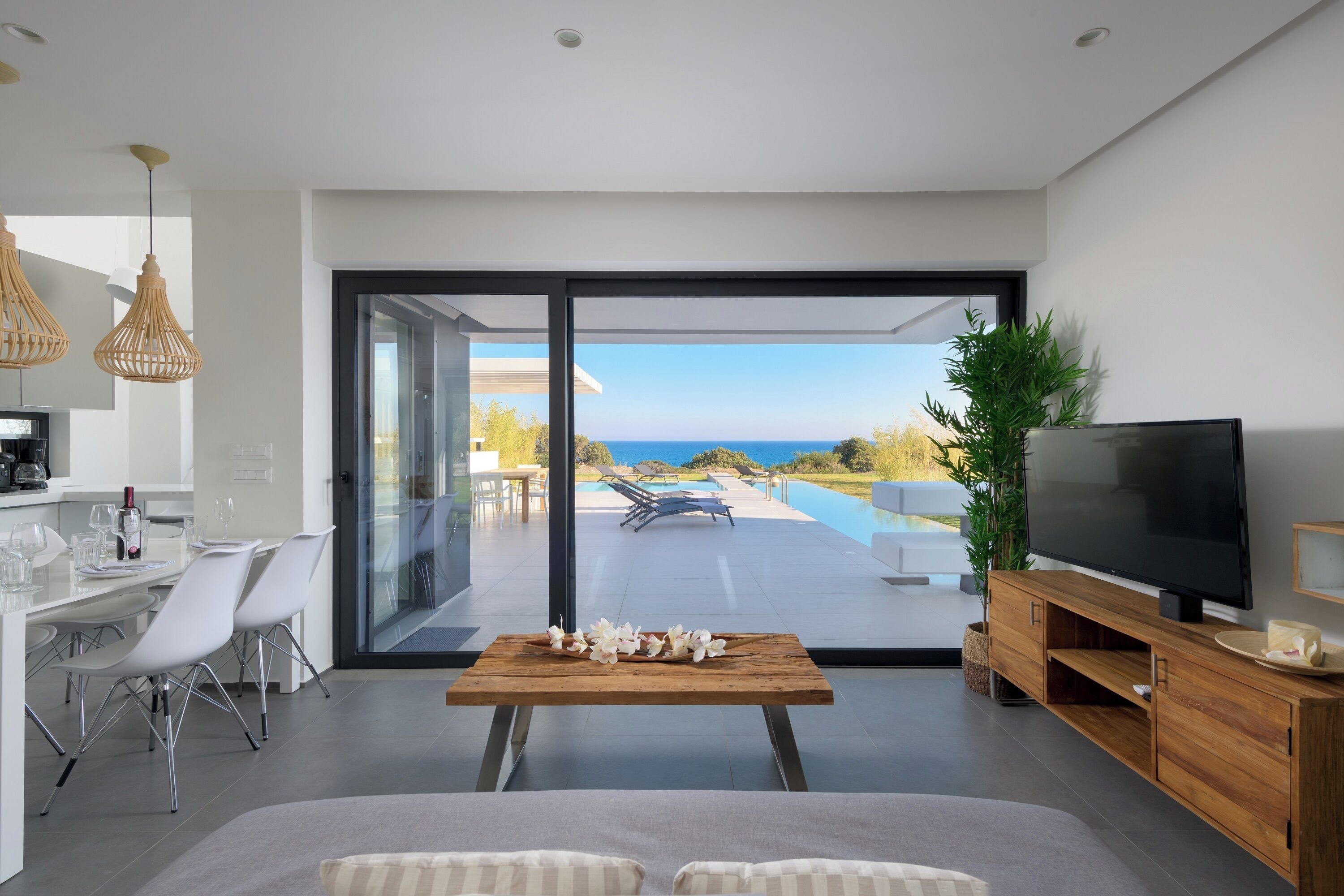 A modern living room with a large TV, a dining area with chairs, a wooden table with flowers, and a view of the ocean through large glass doors.