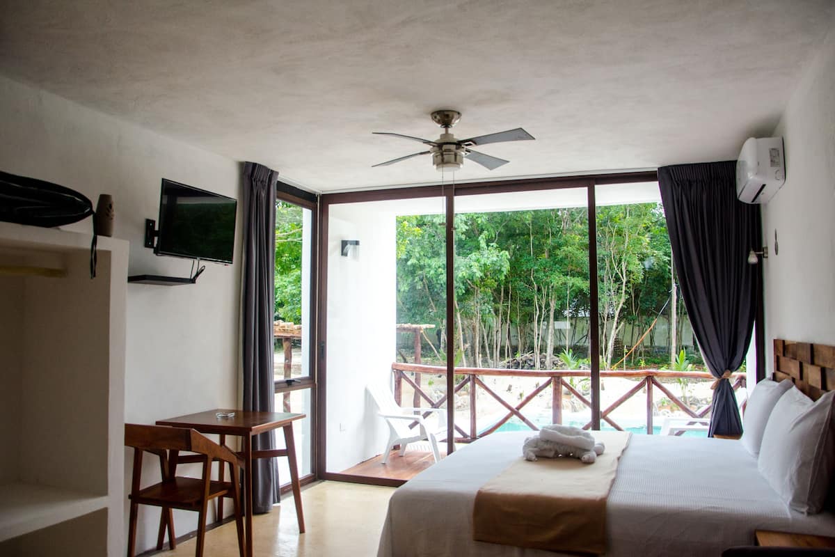 Habitación de hotel con cama, escritorio, silla, televisión y balcón con vistas a árboles.