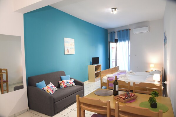 A room with a blue wall, a sofa with colorful pillows, a wooden table with a bottle, and a bed with a nightstand. - Economic Sudios B4 (Iraklio Crete Greece)