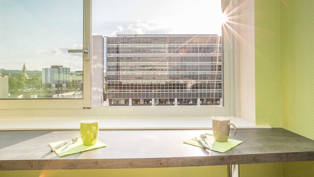 A room with a window overlooking a cityscape, a table with two mugs, and a spoon on a napkin. - Chambre sympa Chambre d'hôtes in the heart of Nancy (Nancy)