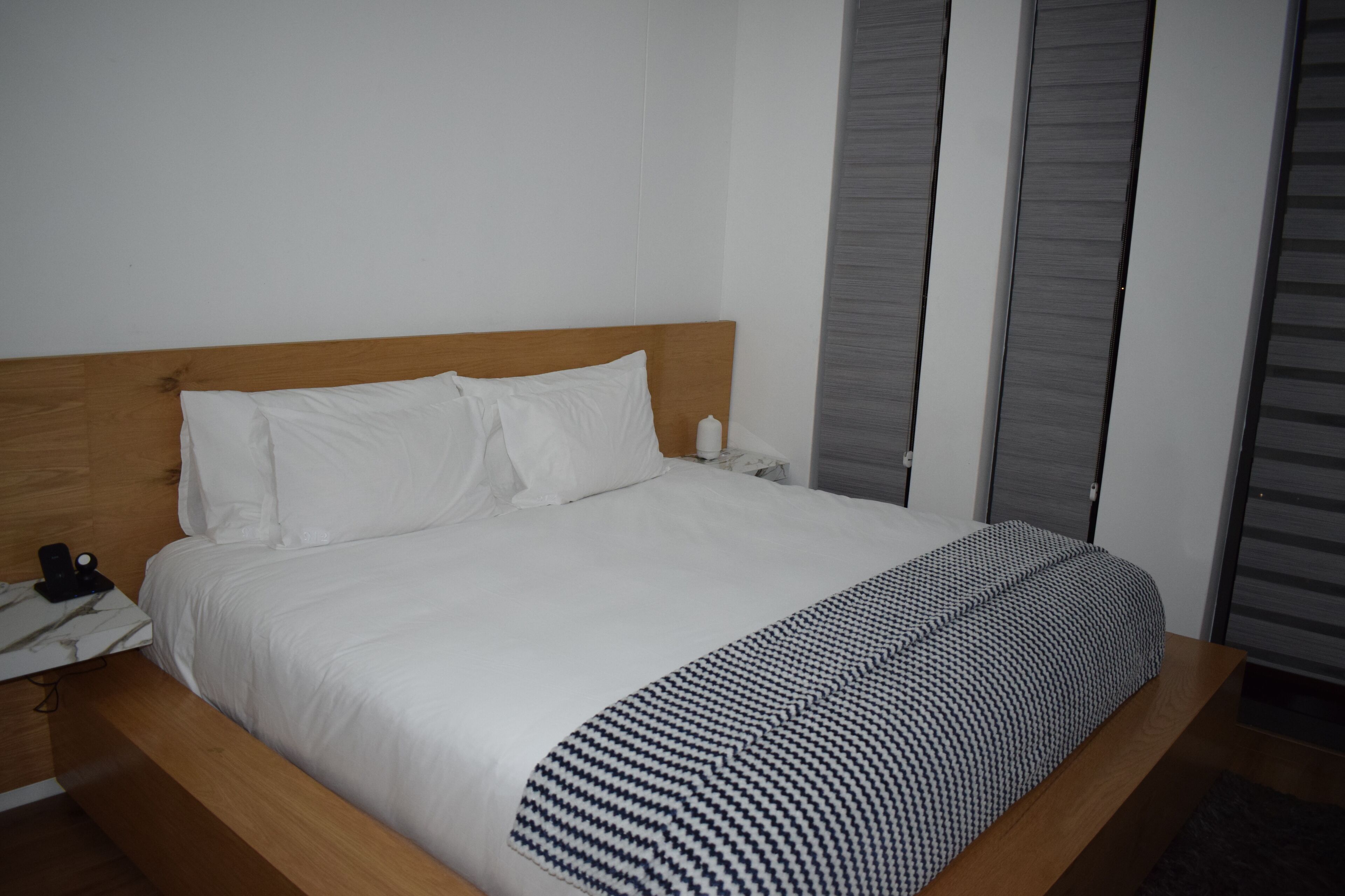 A neatly made bed with white bedding and a patterned throw blanket, a bedside table with a phone, and a wall-mounted light.