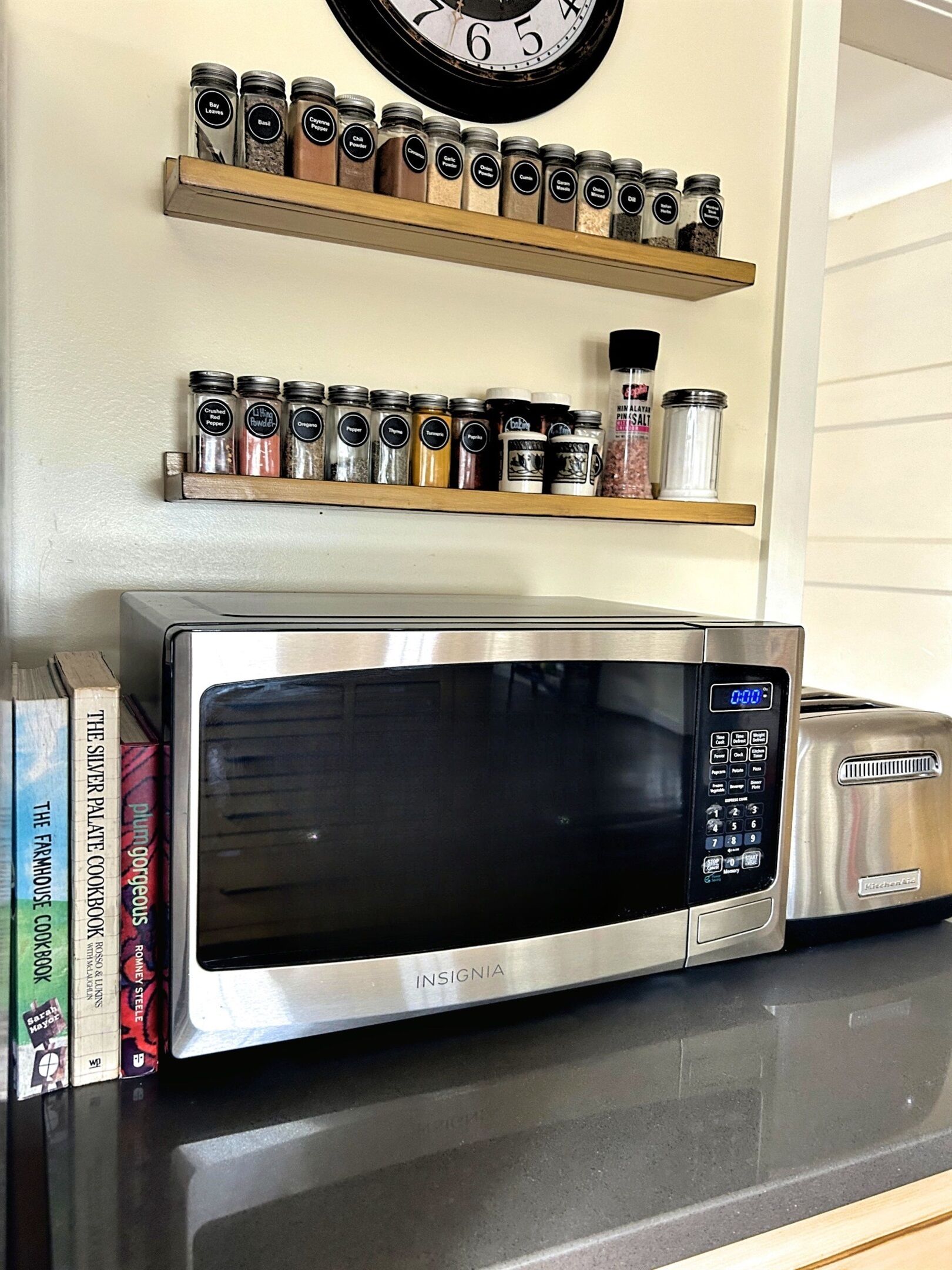 A kitchen with a stainless steel microwave oven, two spice racks, and a clock on the wall.