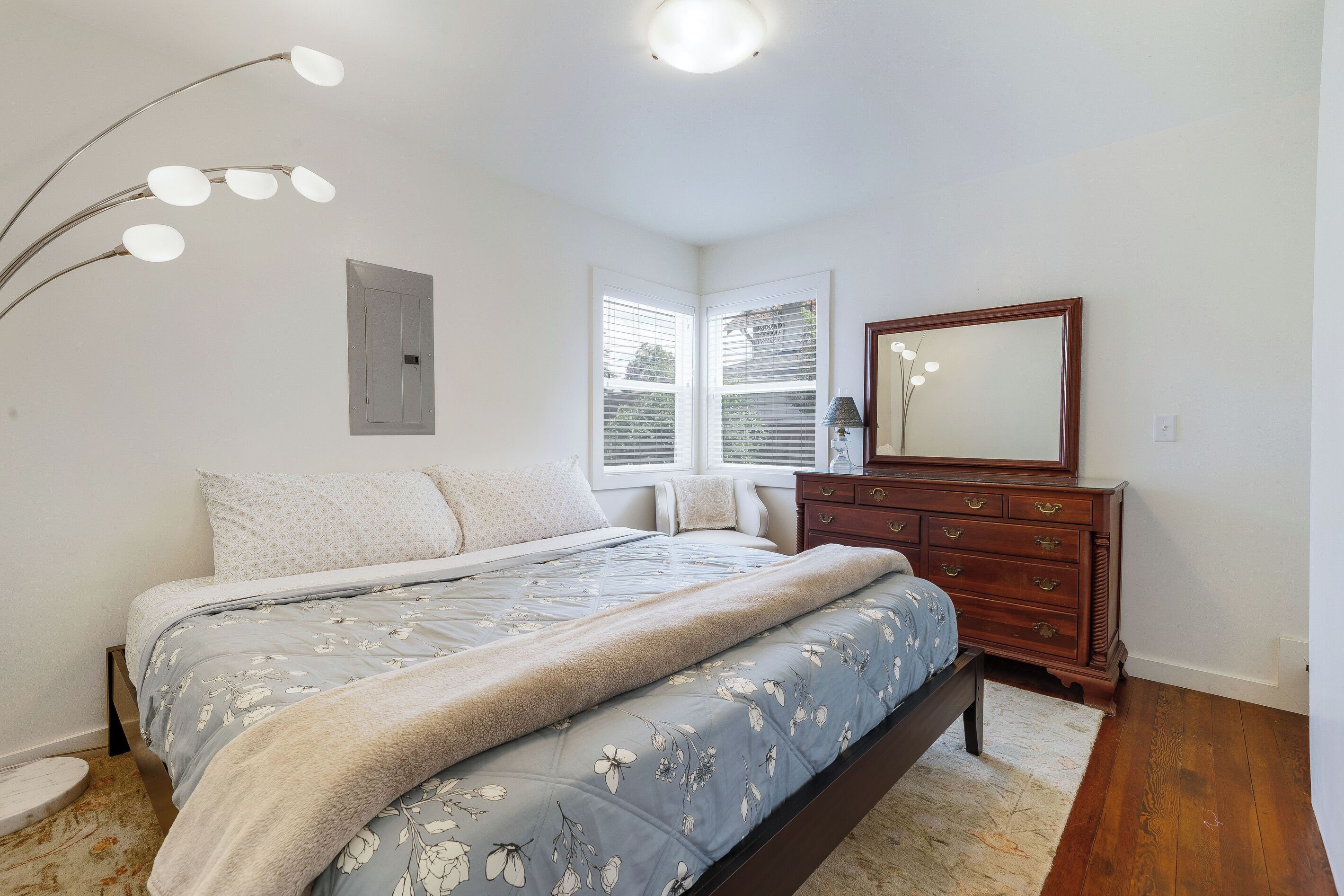 Ein Schlafzimmer mit Bett, Kommode, Spiegel und einem Fenster mit Jalousien.