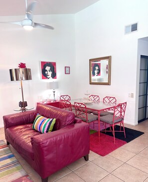 Una sala de estar con un sofá rojo, una mesa de comedor con sillas rojas, un ventilador de techo y decoración en la pared.