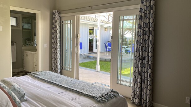 Un dormitorio con cama, un baño con lavamanos y espejo, y un ventanal corredero que da a un patio exterior con sillas y una mesa.