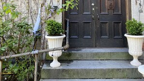 Una puerta doble de madera oscura con un aldabón decorativo, flanqueada por dos urnas blancas con plantas verdes, que da acceso a una escalera de piedra.