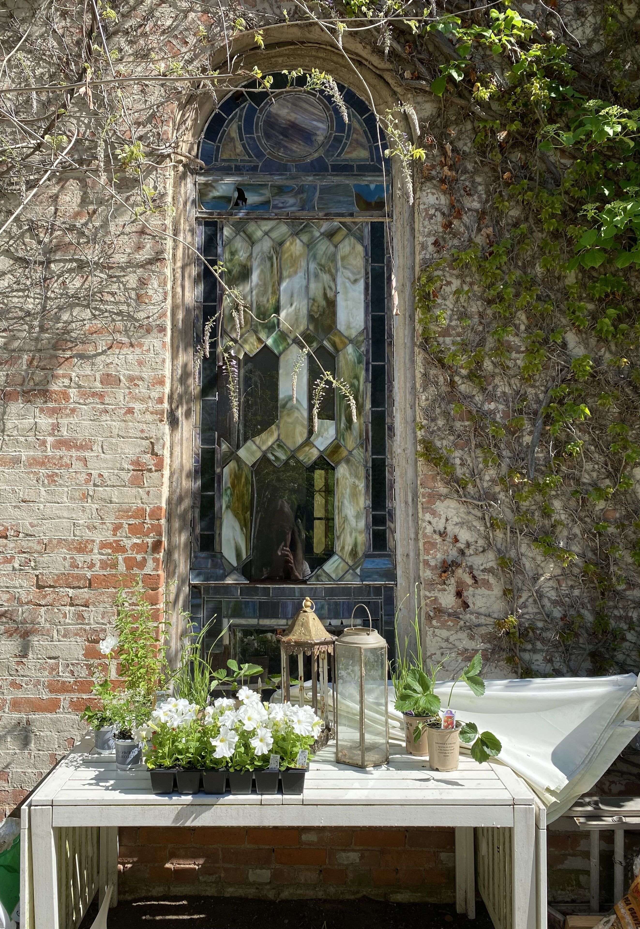 A rustic building with a stained glass window, a brick wall, and a table with potted plants and a lantern.
