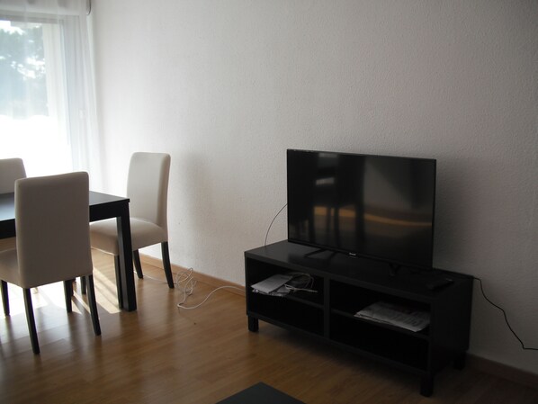 A modern living room with a flat-screen TV on a stand, a dining table with chairs, and a window with curtains.