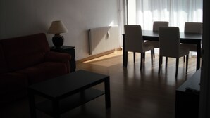 Un salon comprenant un canapé, une table basse, une lampe, une table à manger avec des chaises et une grande fenêtre ornée de rideaux légers.
