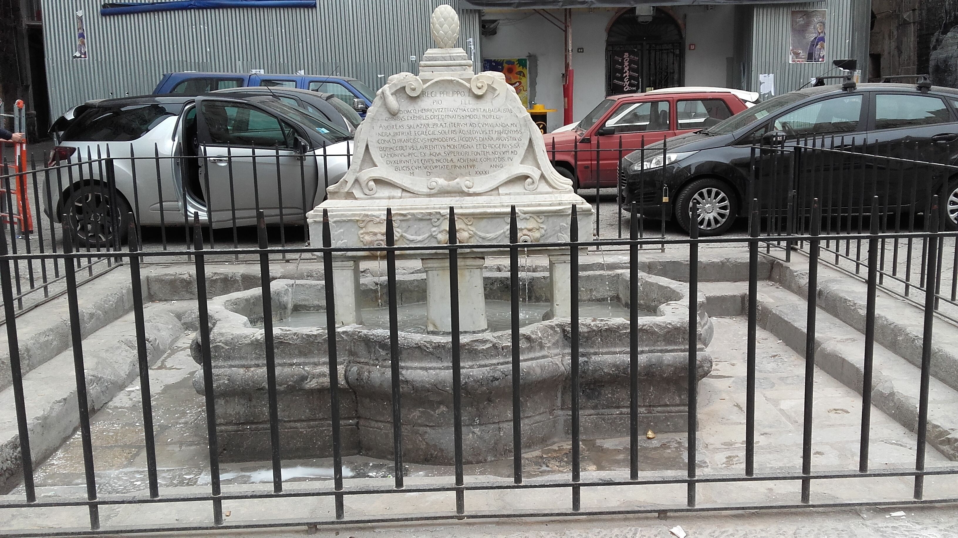 Een stenen fontein met inscripties, omringd door een metalen hek, met auto's in de buurt geparkeerd.
