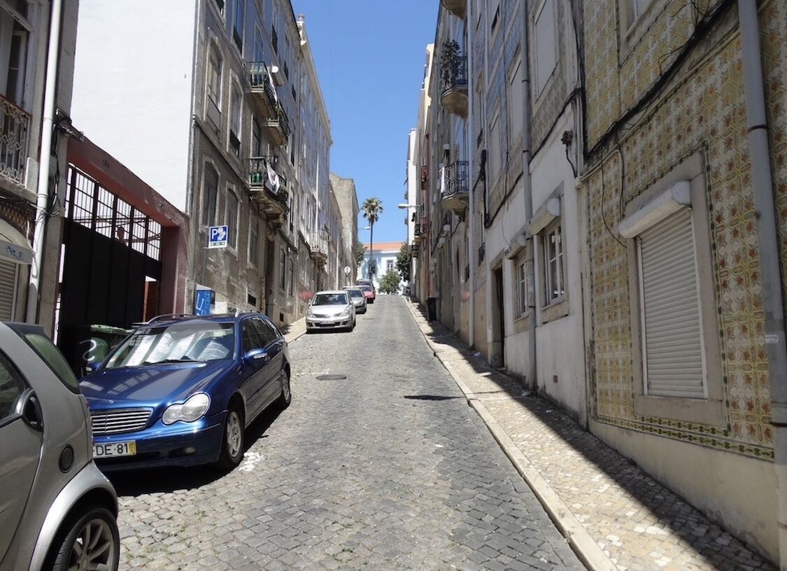 A narrow cobblestone street lined with buildings, parked cars, and a clear blue sky.
