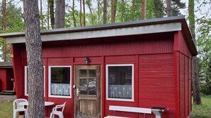 A small red cabin with a wooden door, two windows with curtains, a bench, and a white chair outside.