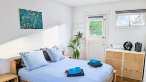 Un dormitorio con una cama de madera, una mesita de noche pequeña, una planta en maceta, un radiador y una ventana con vistas a zonas verdes.