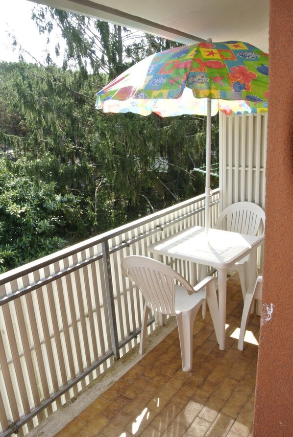 Un balcón con una mesa y sillas blancas, un paraguas colorido y vistas a zonas verdes.