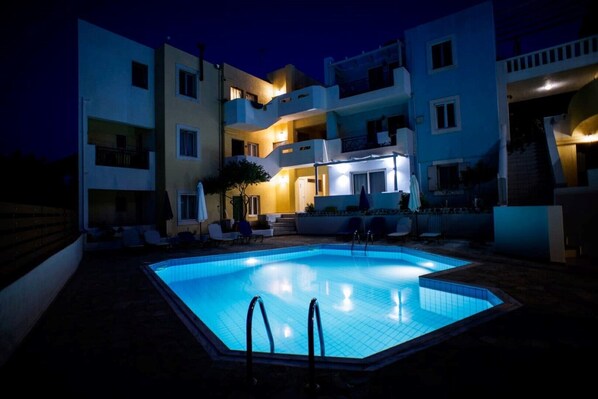 Una piscina con sillones de descanso y una zona de estar techada, rodeada de edificios de varios pisos con balcones.