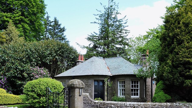 A stone house with a slate roof, surrounded by lush greenery and a stone wall.