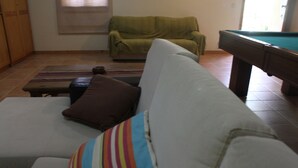Un salon avec un canapé blanc, un fauteuil vert, une table de billard et un coussin à rayures.