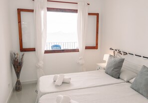 A bedroom with two beds, a window with curtains, a bedside table with a lamp, and a view of a balcony.